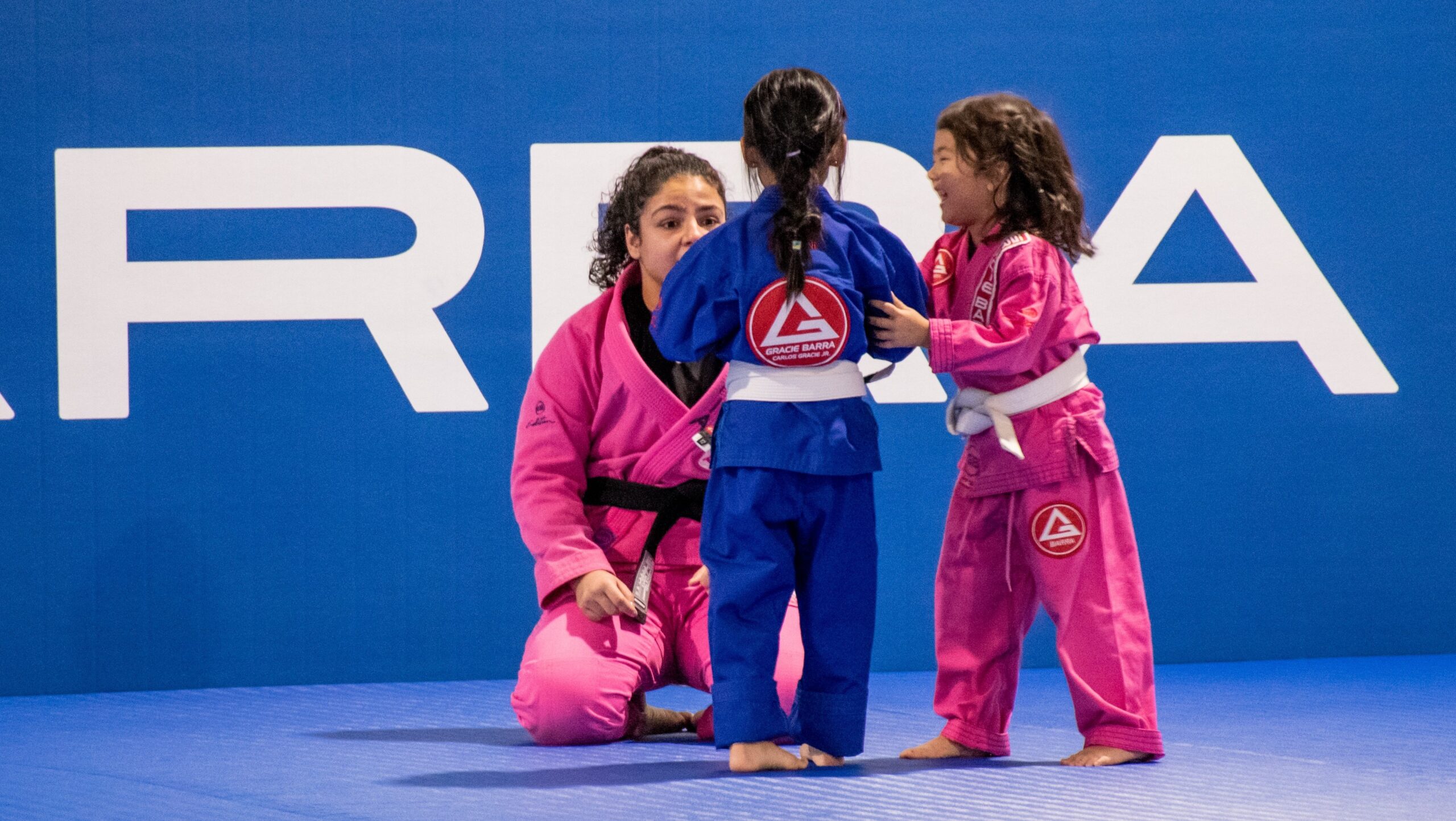 Two young kids in kimonos practicing Jiu-Jitsu guided by an instructor on blue mats.